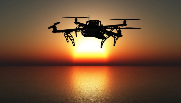The silhouette of a drone with four propellers is shown against a bright orange and yellow sunset over the ocean.